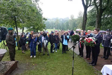 Uczestnicy uroczystości, fotoreporterzy i operatorzy kamer na trawniku przy pomniku; po prawej na alejce delegacje z wieńcami biało-czerwonych kwiatów, na pierwszym planie mikrofon.