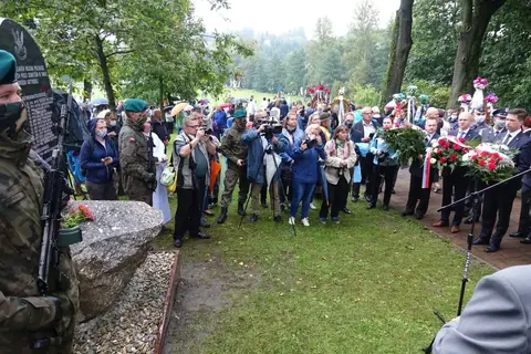 Widok zza żołnierza asysty: kamienny głaz pomnika, fotoreporterzy z kamerami i delegacje z wieńcami na alejce.