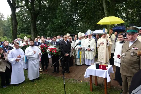 Biskupi w mitrach, księża, ministranci i delegacje z wieńcami przed stolikiem z kropidłem; na pierwszym planie oficer w mundurze.