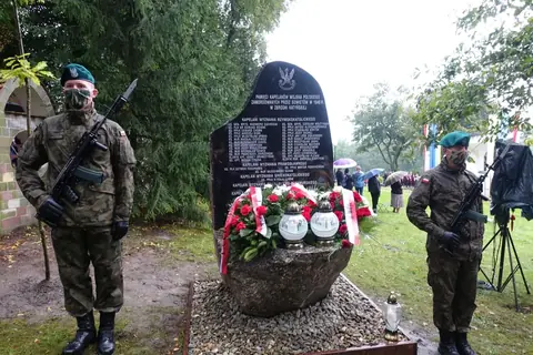 Pomnik z tablicą, wieńcami i zniczami, po bokach żołnierze wojskowej asysty.